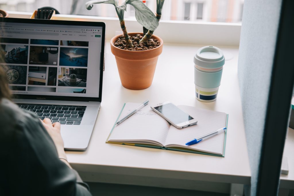 A lady sitting at a desk with her laptop, notebook, phone, and coffee. She is looking out of a window and has a small house plant in front of her.