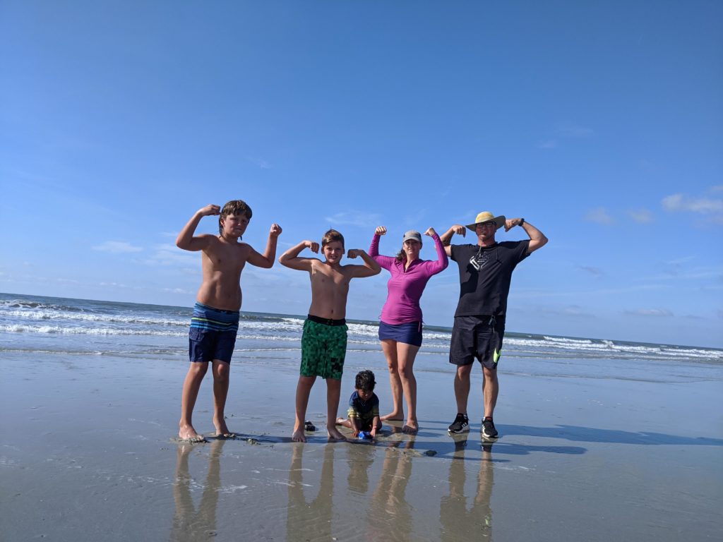 Beach picture with the ocean in the background. Ben Jacqueline and 2 oldest boys are standing in their swimsuits and flexing muscled. The youngest son is sitting and playing in the sand.