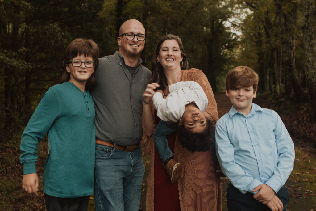 Family picture outdoors with Ben, Jacqueline, and 3 boys. It is the fall and they are surrounded trees and leaves on the ground. It is a silly picture with the 3 year old bent over backwards goofing off.