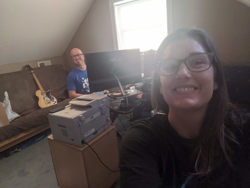 Selfie of Jacqueline with brown hair and glasses, grinning at the camera. She's backlit heavily by a window, partially obscuring her face. Ben (bald with glasses, wearing a blue tshirt) is peeking around a monitor on a desk under the window. Behind him is a brown futon with an electric guitar sitting upright on it.