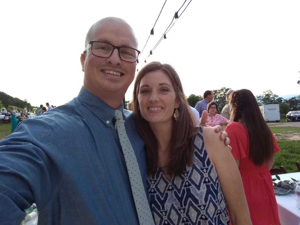 Selfie of Ben and Jacqueline at a n outdoor summer wedding reception. He is wearing a blue shirt and she is wearing a sleeveless blue and grey dress.