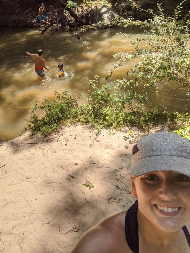 Selfie of Jacqueline wearing a hat with the boys playing in a river behind her.