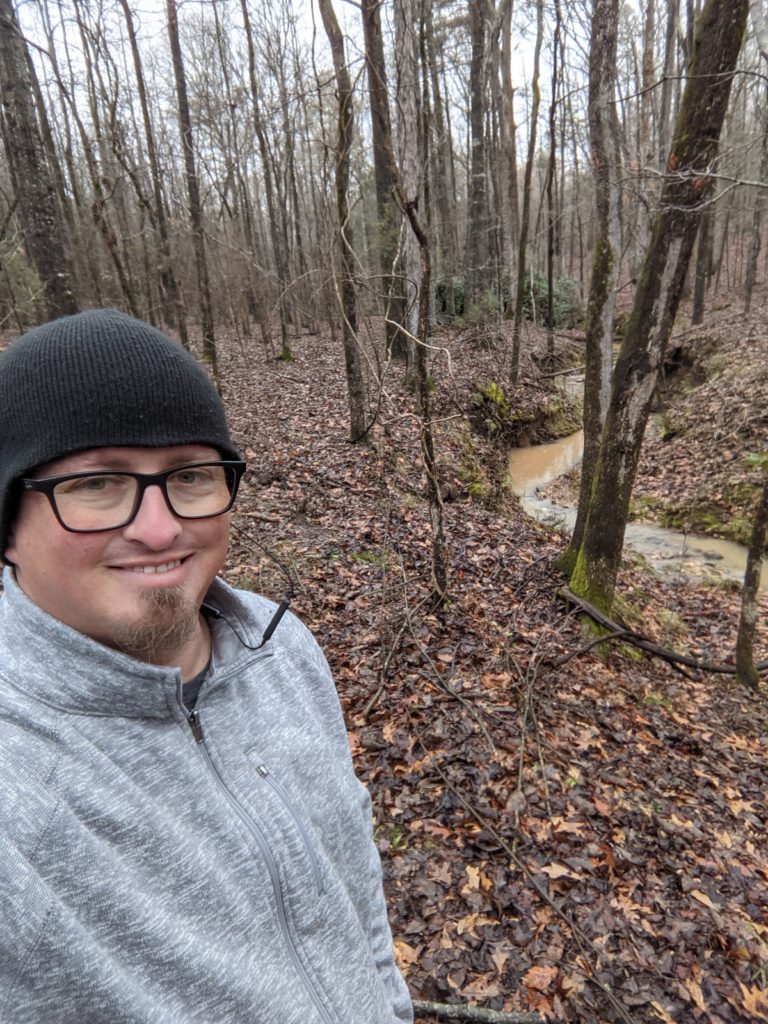 Selfie of Ben in the woods. He is wearing a black toboggan and grey jacket. There are leaves on the ground and a muddy creek behind him.
